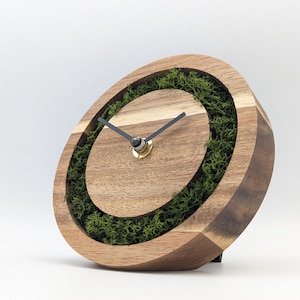 Op de afbeelding: Houten bureauklok met een natuurlijk ontwerp. De klok heeft een lichtbruin houten frame met een cirkelvormige inzet gevuld met groen mos. Zwarte wijzers en een goudkleurig middenstuk maken het ontwerp compleet. De klok is ongeveer 15 cm hoog.