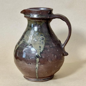 May include: A handcrafted ceramic pitcher featuring a bulbous body and a curved handle. The surface displays a deep brown glaze with mottled green and tan drip patterns cascading down the side, creating a textured, glossy finish with a spout.