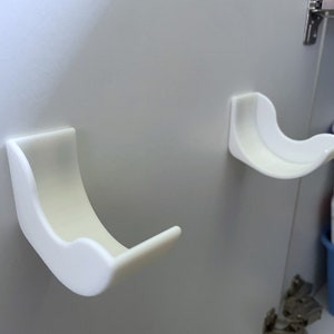 Può includere: Due ganci in plastica bianca curva sono montati su una superficie grigio chiaro. I ganci hanno una finitura liscia e opaca e sono progettati con una forma arrotondata. I ganci sono probabilmente per appendere oggetti.