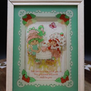 May include: A framed vintage illustration featuring cartoon characters around a table. The frame is white with a green border and strawberry accents. The image includes the text "Add heaven's love to all you do And goodness will come back to you."