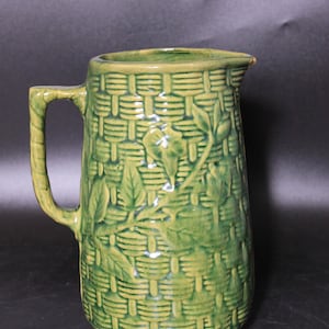 May include: A green ceramic pitcher featuring a raised basket weave texture across the exterior surface. The pitcher is decorated with embossed leaf and vine patterns and includes a single handle on the side and a pouring spout at the top rim.