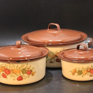 May include: A set of three brown and cream enameled metal cooking pots with lids. Each pot features a decorative band of colorful vegetable illustrations including tomatoes, peppers, and artichokes around the exterior base of the cookware.