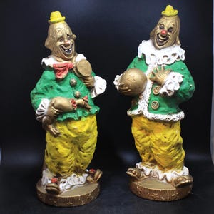 May include: Two clown figurines with gold-painted faces, yellow hats, and green jackets. The clowns wear yellow pants and white ruffled collars. One holds a mirror, the other a gold ball. The figurines stand on gold bases.