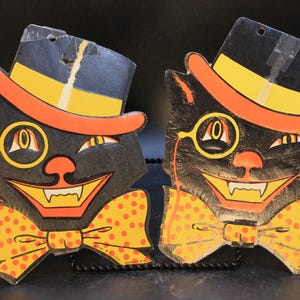 May include: Two identical die-cut cardboard decorations featuring a black cat face wearing a top hat with an orange band and a yellow bow tie with orange polka dots. The cat has a monocle over one eye, a wide grin with visible fangs, and a nose.