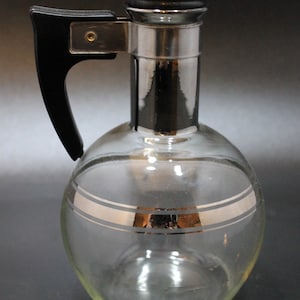 May include: A clear glass decanter with a silver-colored top and a black handle. The decanter has a silver band around the middle and a silver band around the top.