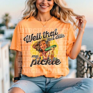 Pu&ograve; includere: Una donna indossa una maglietta arancione con una grafica vintage di una donna che tiene un cetriolo. Il testo sulla maglietta recita "Well that just dills my pickle".