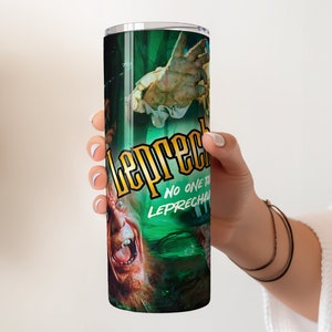 May include: A stainless steel tumbler with a green, black, and yellow design featuring a character from the film "Leprechaun" with the text "Leprechaun" and "No one tricks the Leprechaun".