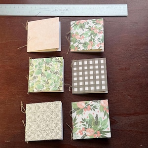 May include: Six small, square notebooks with decorative covers, each bound with twine. The covers feature various patterns, including floral, leaf, and geometric designs. A ruler is positioned above the notebooks on a dark wood surface.