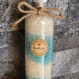 Puede incluir: Una botella de vidrio transparente llena de sales de baño blancas y azules, atada con una cuerda y un lazo. La botella tiene una etiqueta que dice "Hand Made With Love - thank you - Especially For You".