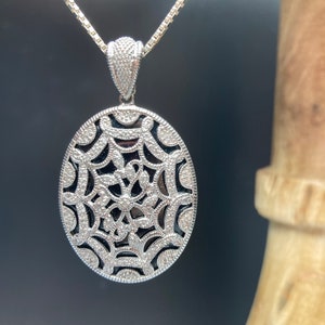 May include: A silver pendant necklace with a detailed oval-shaped design featuring a black spiderweb pattern. The pendant is set with small clear stones.