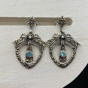 May include: A pair of silver earrings with a blue stone in the center of each earring. The earrings are made of a delicate, intricate design with a floral motif.