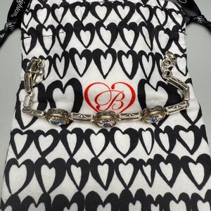 Puede incluir: Una pulsera plateada con tres piedras preciosas ovaladas y transparentes engastadas en biseles dorados. La pulsera descansa sobre una bolsa blanca con cord&oacute;n y estampado de corazones negros. La bolsa tiene un coraz&oacute;n rojo con un logotipo "B" estilizado.