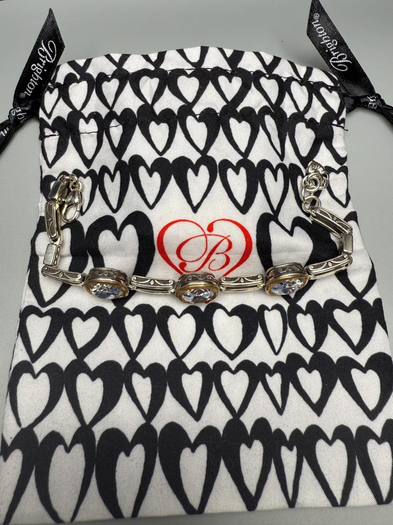 Puede incluir: Una pulsera plateada con tres piedras preciosas ovaladas y transparentes engastadas en biseles dorados. La pulsera descansa sobre una bolsa blanca con cord&oacute;n y estampado de corazones negros. La bolsa tiene un coraz&oacute;n rojo con un logotipo "B" estilizado.