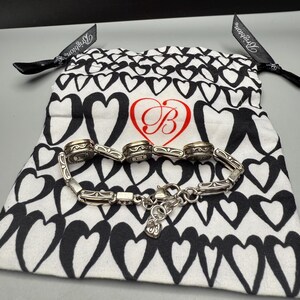 Puede incluir: Una pulsera plateada con eslabones decorativos y un cierre de mosquet&oacute;n, colocada sobre una bolsa blanca con cord&oacute;n y estampado de corazones negros. La bolsa presenta un coraz&oacute;n rojo con la letra estilizada "B". Cintas negras est&aacute;n unidas a la bolsa.