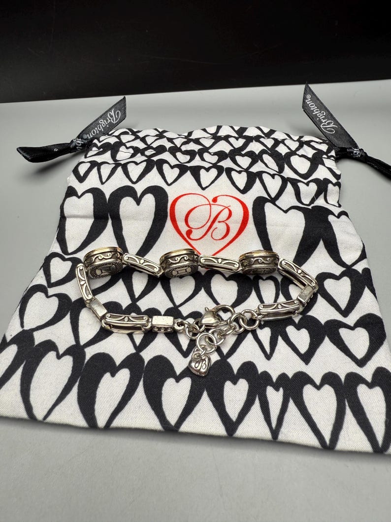 Puede incluir: Una pulsera plateada con eslabones decorativos y un cierre de mosquet&oacute;n, colocada sobre una bolsa blanca con cord&oacute;n y estampado de corazones negros. La bolsa presenta un coraz&oacute;n rojo con la letra estilizada "B". Cintas negras est&aacute;n unidas a la bolsa.