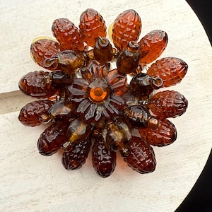 May include: A large amber glass brooch with a central flower design and a clear rhinestone in the center. The brooch is made up of many small, teardrop-shaped glass beads.