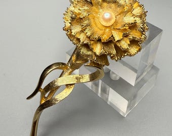 May include: A gold-toned brooch in the shape of a carnation with a single pearl in the center. The flower is detailed with textured petals and a stem with a twisted design.