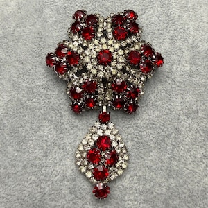 May include: A vintage brooch with a starburst design, featuring ruby red and clear crystal rhinestones. The brooch has a pendant with a teardrop shape, also adorned with red and clear crystals. The setting is silver-toned.