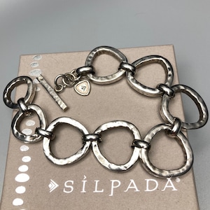 May include: Silver-toned bracelet with hammered oval links and a toggle clasp. The bracelet is resting on a cardboard box with the brand name "SILPADA" printed on it. The bracelet has a small heart-shaped charm.