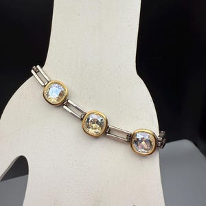 Puede incluir: Pulsera plateada con tres cristales grandes, cuadrados y transparentes engastados en biseles dorados. La pulsera se exhibe sobre una forma blanca con forma de mano sobre un fondo negro. Los cristales tienen un ligero tinte amarillo.