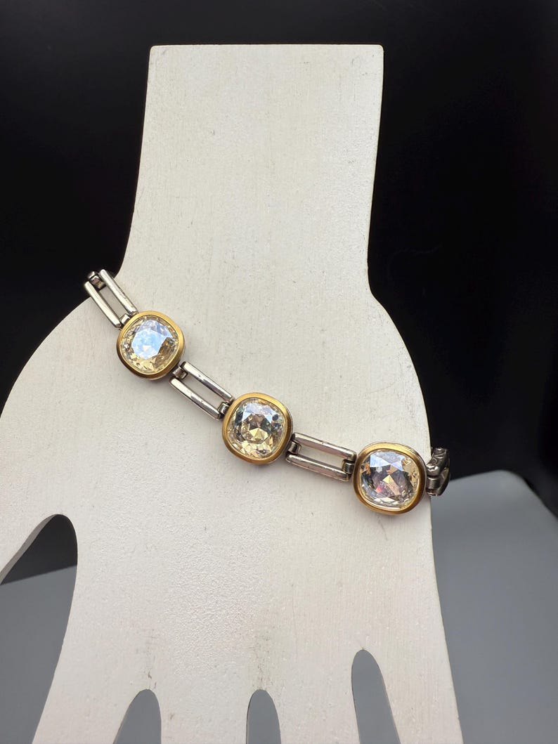 Puede incluir: Pulsera plateada con tres cristales grandes, cuadrados y transparentes engastados en biseles dorados. La pulsera se exhibe sobre una forma blanca con forma de mano sobre un fondo negro. Los cristales tienen un ligero tinte amarillo.