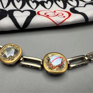 Puede incluir: Un brazalete plateado con biseles cuadrados dorados que sostienen piedras claras y facetadas. El brazalete est&aacute; sobre una superficie gris, con una tela con estampado de corazones en blanco y negro en el fondo.