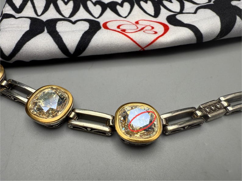 Puede incluir: Un brazalete plateado con biseles cuadrados dorados que sostienen piedras claras y facetadas. El brazalete est&aacute; sobre una superficie gris, con una tela con estampado de corazones en blanco y negro en el fondo.