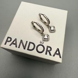 May include: A pair of silver hoop earrings with a dangling square charm, both adorned with clear cubic zirconia stones. The earrings are displayed on a white box with the word "PANDORA" printed in black.