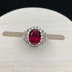 May include: A silver ring with a large oval-shaped red gemstone surrounded by smaller clear gemstones.