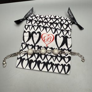 Puede incluir: Una pulsera plateada con tres piezas centrales cuadradas y texturizadas. La pulsera se exhibe sobre una bolsa blanca con cord&oacute;n y estampado de corazones negros y un logotipo "B" rojo. Cintas negras con texto "Brighton" atadas a la bolsa.