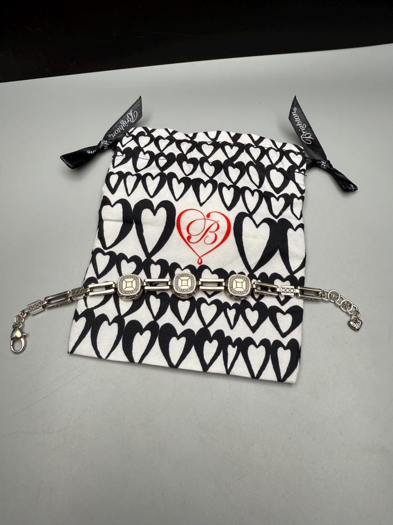 Puede incluir: Una pulsera plateada con tres piezas centrales cuadradas y texturizadas. La pulsera se exhibe sobre una bolsa blanca con cord&oacute;n y estampado de corazones negros y un logotipo "B" rojo. Cintas negras con texto "Brighton" atadas a la bolsa.
