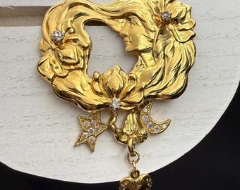 May include: A gold-toned brooch featuring a woman's profile within a floral wreath design. The brooch has a dangling charm with a heart, star, and crescent moon, all adorned with sparkling crystals. The brooch is a vintage-inspired accessory.