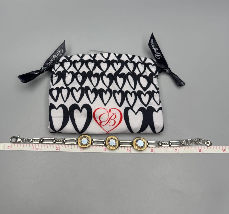 Puede incluir: Una pulsera plateada con tres piezas centrales cuadradas, doradas y opalescentes. Una bolsa blanca con corazones negros y un coraz&oacute;n rojo con un logotipo "B" est&aacute; detr&aacute;s de la pulsera. La pulsera mide aproximadamente 18 cm de largo.