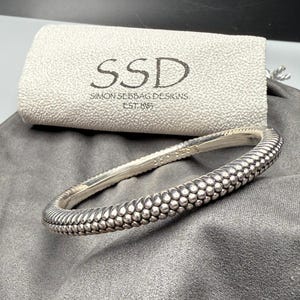 May include: A silver bracelet with a textured, beaded design. The bracelet is next to a light gray pouch with the text "SSD SIMON SEBBAG DESIGNS EST. 1985". The bracelet has a curved shape and a detailed pattern.