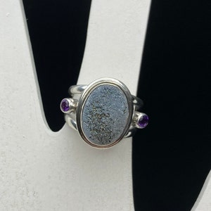 May include: A silver ring with a gray oval stone set in a bezel setting. The stone is surrounded by a silver band with two small purple stones on either side.