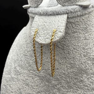 May include: A pair of gold-toned rope chain earrings. Each earring features a continuous, twisted metal link design that forms a slender, elongated loop. The earrings are suspended from a textured, light gray display stand with a soft surface.
