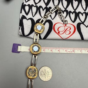 Puede incluir: Una pulsera plateada y dorada con tres piedras cuadradas e iridiscentes. La pulsera est&aacute; junto a una bolsa con estampado de corazones en blanco y negro y una moneda de diez centavos. Una cinta m&eacute;trica muestra cent&iacute;metros.