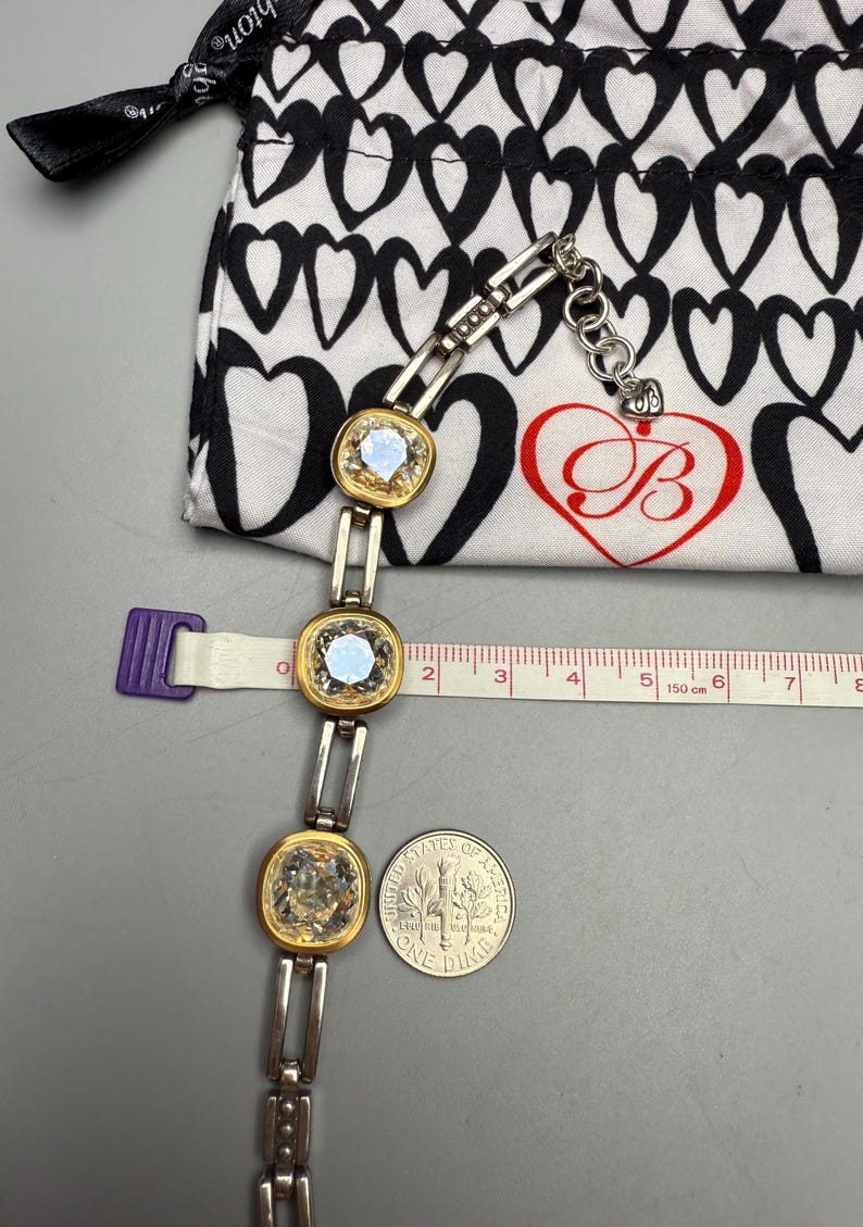 Puede incluir: Una pulsera plateada y dorada con tres piedras cuadradas e iridiscentes. La pulsera est&aacute; junto a una bolsa con estampado de corazones en blanco y negro y una moneda de diez centavos. Una cinta m&eacute;trica muestra cent&iacute;metros.