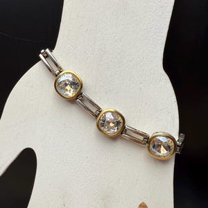 Puede incluir: Un brazalete plateado y dorado con tres cristales grandes y transparentes de corte cuadrado. Los cristales est&aacute;n engastados en biseles dorados y conectados por eslabones plateados. El brazalete se muestra sobre una superficie blanca sobre un fondo negro.