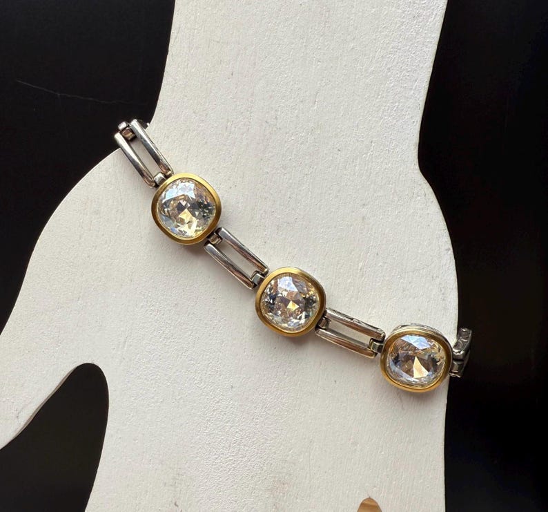 Puede incluir: Un brazalete plateado y dorado con tres cristales grandes y transparentes de corte cuadrado. Los cristales est&aacute;n engastados en biseles dorados y conectados por eslabones plateados. El brazalete se muestra sobre una superficie blanca sobre un fondo negro.