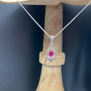 May include: A silver chain necklace with a pink gemstone pendant. The pendant is shaped like a teardrop and is surrounded by smaller clear gemstones.
