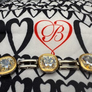 Puede incluir: Una pulsera plateada y dorada con tres gemas claras grandes, de talla cuadrada. Las gemas est&aacute;n engastadas en biseles dorados. El fondo es una tela con estampado de corazones en blanco y negro con un coraz&oacute;n rojo y la letra "B".