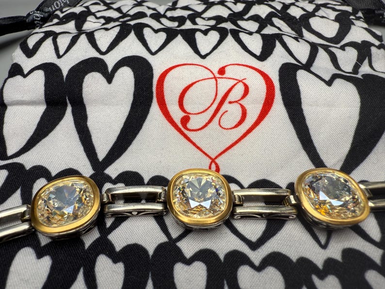 Puede incluir: Una pulsera plateada y dorada con tres gemas claras grandes, de talla cuadrada. Las gemas est&aacute;n engastadas en biseles dorados. El fondo es una tela con estampado de corazones en blanco y negro con un coraz&oacute;n rojo y la letra "B".