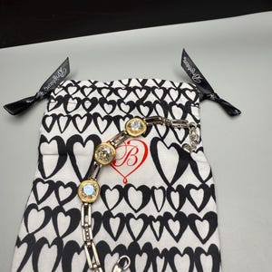 Puede incluir: Una pulsera plateada con tres piedras de cristal transparente grandes y redondas engastadas en biseles dorados. La pulsera se muestra sobre una bolsa blanca con cord&oacute;n con un patr&oacute;n de corazones negros y un coraz&oacute;n rojo con una "B" en su interior. Cintas negras con texto blanco son visibles.