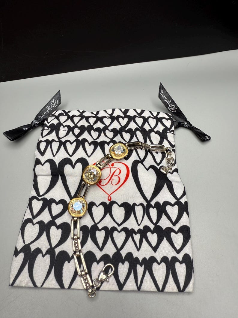 Puede incluir: Una pulsera plateada con tres piedras de cristal transparente grandes y redondas engastadas en biseles dorados. La pulsera se muestra sobre una bolsa blanca con cord&oacute;n con un patr&oacute;n de corazones negros y un coraz&oacute;n rojo con una "B" en su interior. Cintas negras con texto blanco son visibles.