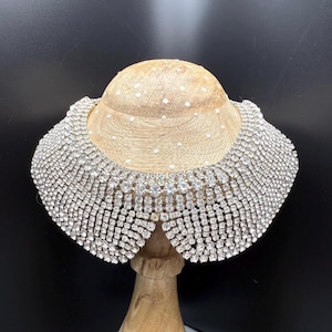 May include: A sparkling, silver rhinestone collar necklace. The collar is displayed on a wooden bust, showcasing its intricate design and brilliant shine. The collar is made of rows of small, square-cut rhinestones.