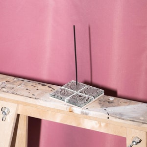May include: A black incense stick stands upright in a square glass incense holder with a swirling black and white pattern. The holder is sitting on a wooden surface.