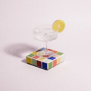 May include: A clear glass cocktail glass with a lime wedge on the rim, sitting on a colorful square tile coaster.