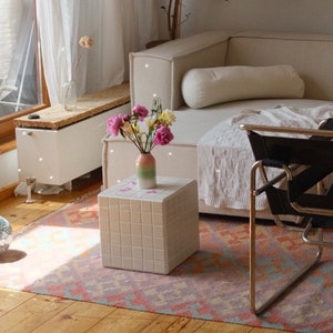 May include: A white, square side table with a grid pattern sits on a pink and orange patterned rug in a living room. A white sofa with a white throw blanket and a black chair with a gold frame are in the background. A vase with pink and yellow flowers sits on the table.