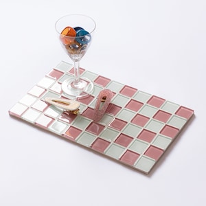 May include: A glass filled with orange and blue hair accessories sits on a pink, white, and green checkered tile. Two hair clips, one gold and one pink, are on the tile. The background is white.